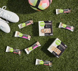Soccer cleats, soccer ball, and ZonePerfect Macros bars sitting on a soccer field